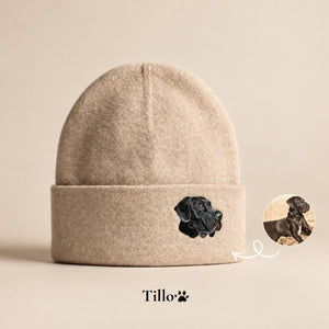 A beige knit beanie with a folded cuff featuring an embroidered black dog head. A small circular inset shows the real dog used for the embroidery, with a white arrow pointing to it.