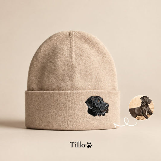 A beige knit beanie with a folded cuff featuring an embroidered black dog head. A small circular inset shows the real dog used for the embroidery, with a white arrow pointing to it.