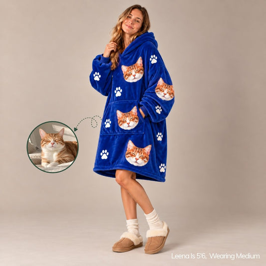 A woman models a royal blue plush hooded robe with a cat-face and paw print pattern, shown against a plain beige studio background. A circular inset shows a cat, with text at the bottom reading 'Leena is 56, Wearing Medium'.