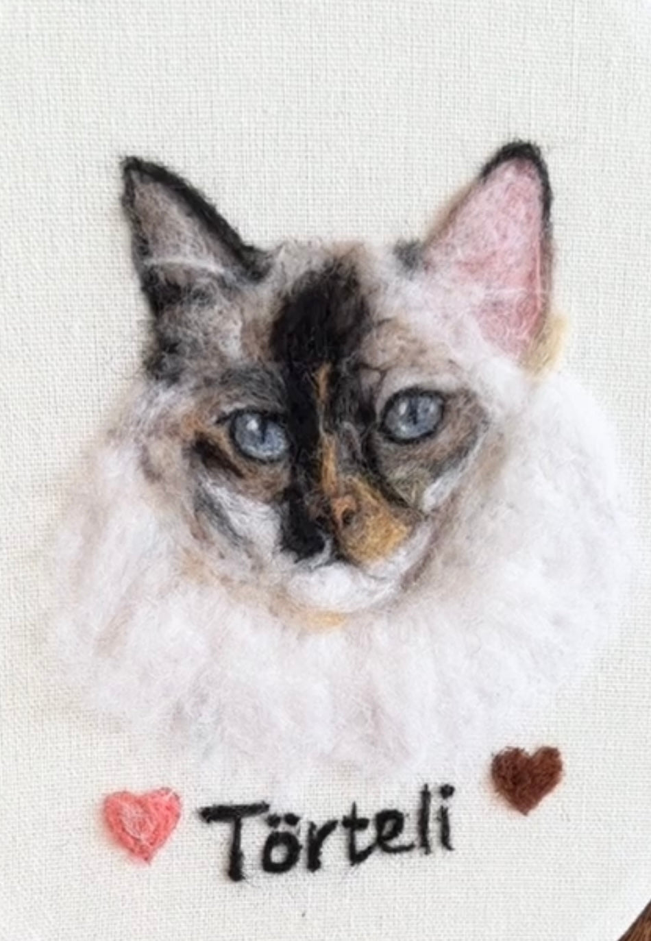 A close-up of a handmade felted wool art piece depicting a cat named Törteli, with two small hearts and the name stitched below on a white felted wool background.